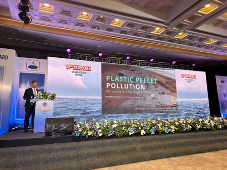 ITOPF staff member on stage presenting on plastic pellets, large screen behind presenter with ITOPF presentation slides, ITOPF presenter wears a dark suit, white shirt and blue tie