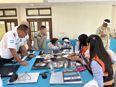 A table-top exercise with maritime stakeholders standing around the table, in the background, someone is using ITOPF's mixed reality application with a headset on